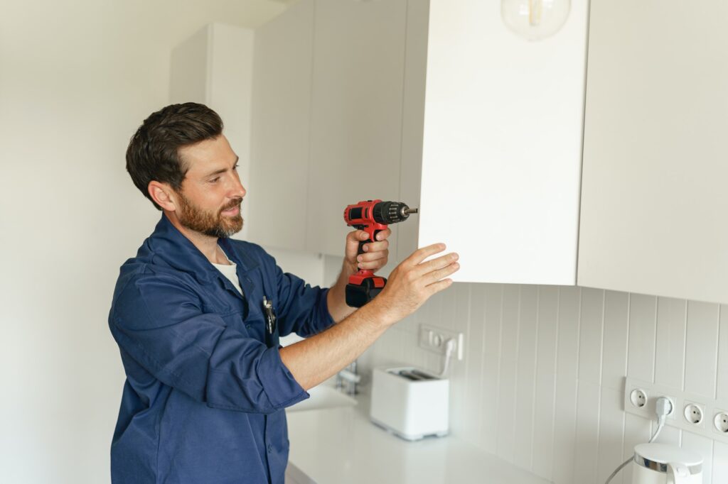 Professional repairman in uniform is using a screwdriver to adjust door of cabinet in kitchen